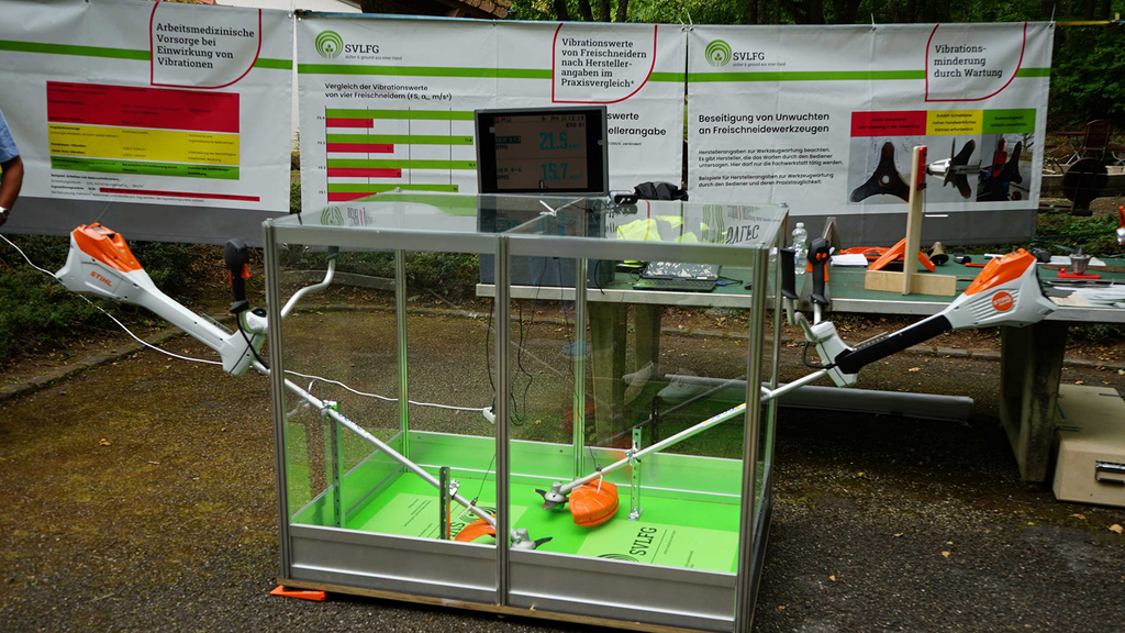 Vergrößerung des Bildes für Ein Ausstellungsstand der SVLFG. Im Hintergrund Plakate zu Freischneidern und Vibrationen. Im Vordergrund ein Glaskasten, aus dem zwei Freischneider herausragen und in dem die Vibrationen gemessen werden..