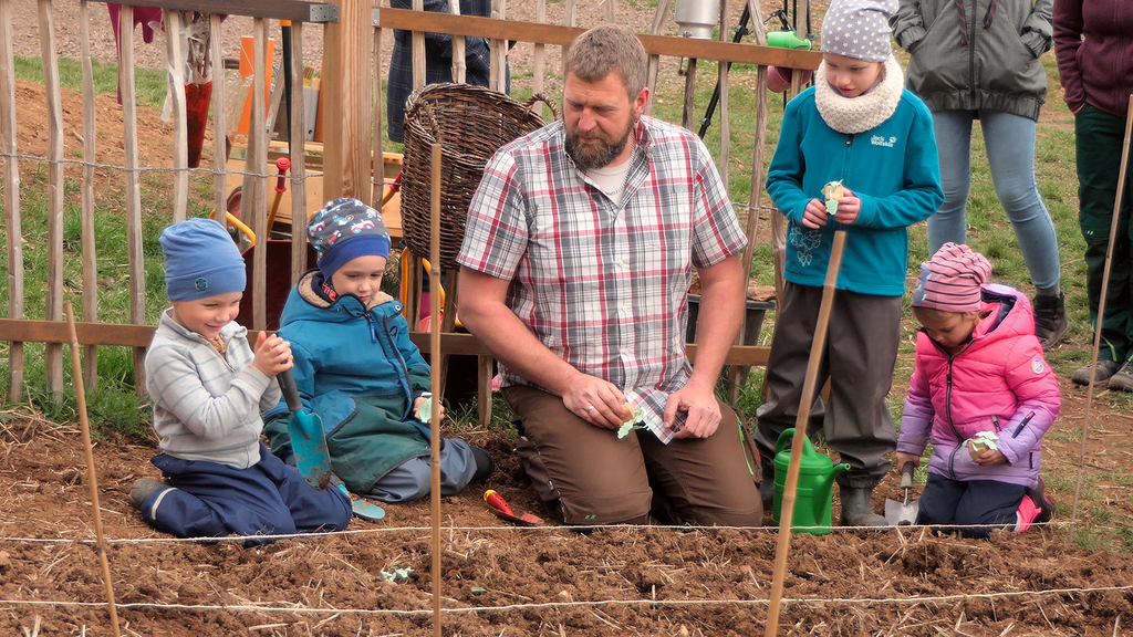 Vergrößerung des Bildes für Vier Kinder und ein erwachsener Mann knien auf einem Feld und setzen Saatgut ein.