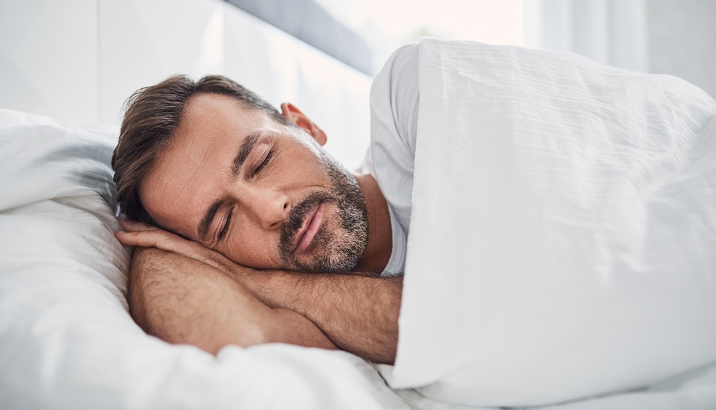 Vergrößerung des Bildes für Mann mittleren Alters liegt mit geschlossenen Augen auf der Seite im Bett. Das Bett ist mit weißer Bettwäsche bezogen. .