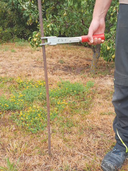 Vergrößerung des Bildes für Eine Hand hält ein Werkzeug, welches eine schnale eiserne Stange umklammert. Die Stange ist im Boden verankert..