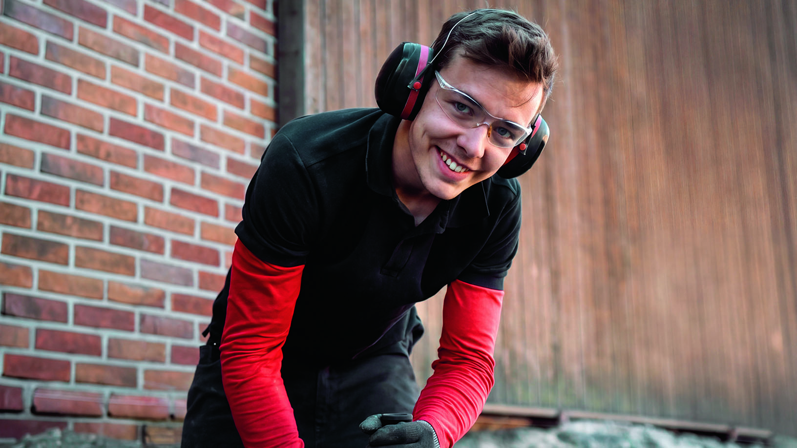Ein junger Mann mit Schutzbrille, Gehörschutz, Arbeitshandschuhen, Arbeitshose steht gebeugt über einem Tisch. Dabei schaut er lächelnd auf und in die Kamera.