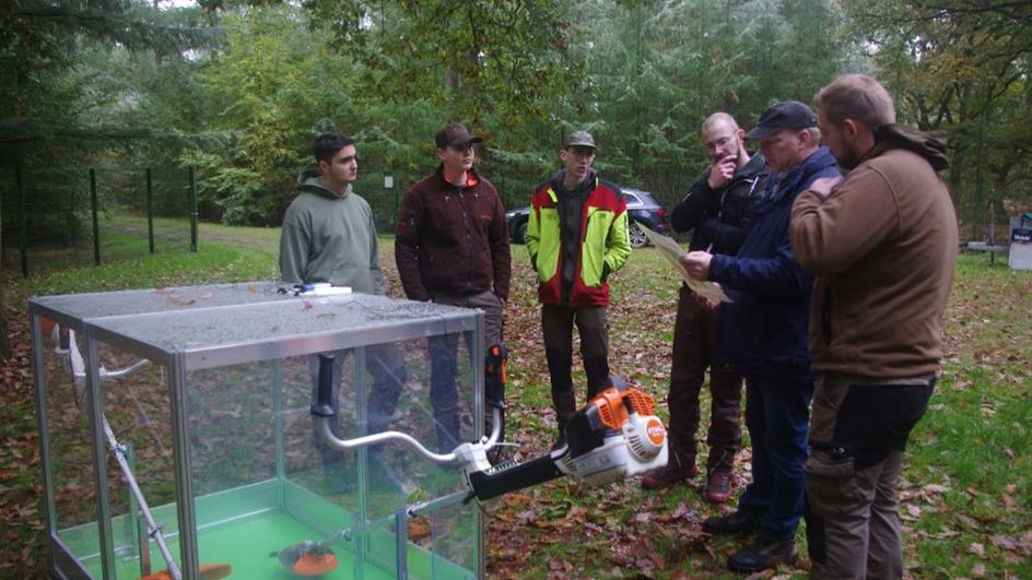 Vergrößerung des Bildes für Mehrere Personen stehen im Wald beisammen. Einer hält ein Papier in der Hand und liest vor. Im Vordergrund ein Freischneidegerät, welches zur Hälfte in einem durchsichtigen Kasten steckt..
