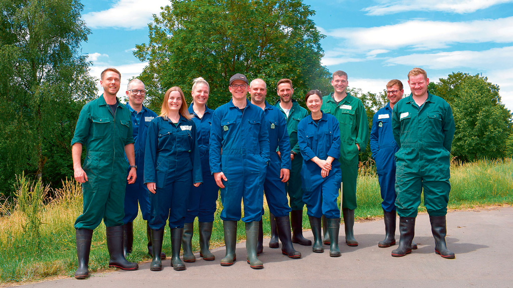 Vergrößerung des Bildes für Gruppenfoto: Mehrere Frauen und Männer und blauen und grünen Overalls und Gummistiefeln stehen draußen und ländlicher Umgebung und lächeln in die Kamera..