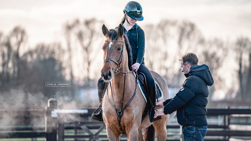 Vergrößerung des Bildes für Eine junge Frau sitzt auf einem Pferd. Ihr Ausbilder steht neben ihr und erklärt ihr wie sie richtig sitzt..