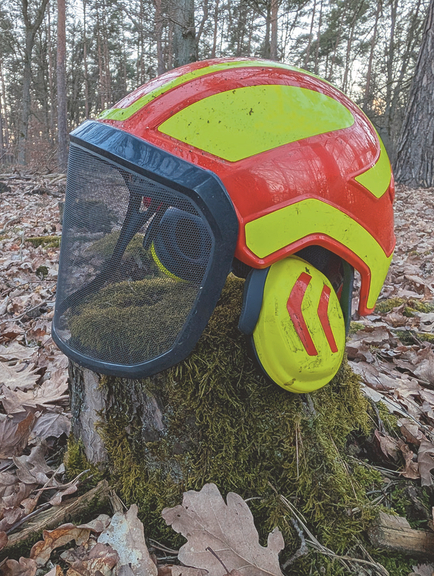 Vergrößerung des Bildes für Ein rot, gelber Schutzhelm mit Visier steht auf einem Baumstumpf. Ohrschützer liegen ebenfalls dabei..