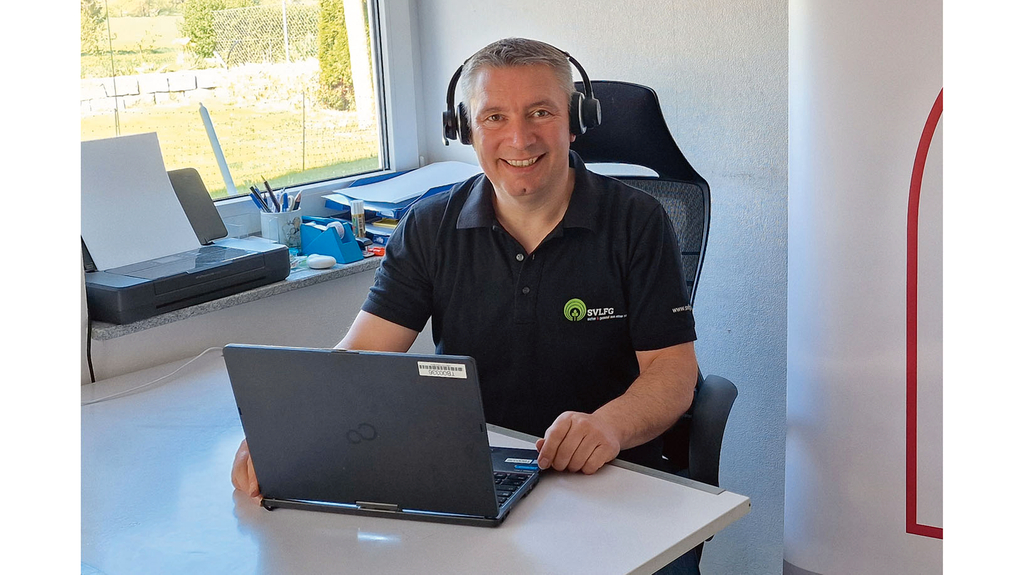 Vergrößerung des Bildes für Ein Mann mit Headset sitzt an einem Schreibtisch vor einem Laptop und schaut freundlich in die Kamera.