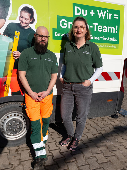 Vergrößerung des Bildes für zwei Personen stehen in Arbeitskleidung der Gärtner vor einem Plakat und schauen in die Kamera..