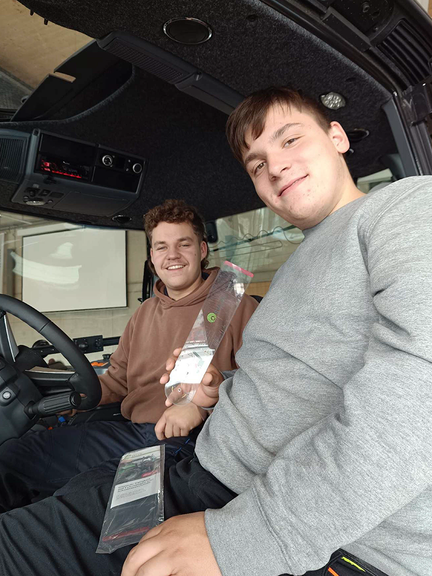 Vergrößerung des Bildes für Zwei junge Männer sitzen in einer Fahrerkabine, halten eine Schablone in der Hand und schauen in die Kamera..