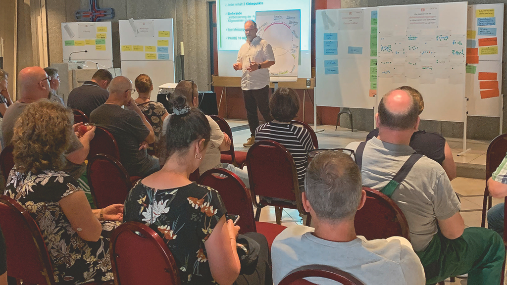 Vergrößerung des Bildes für Abgebildet ist eine Vortragssituation. Der Referent steht vorn und redet. Hinter ihm Flipcharts und Beamer. Vor ihm sitzen mehrere Teilnehmer, teilweise in Arbeitskleidung..