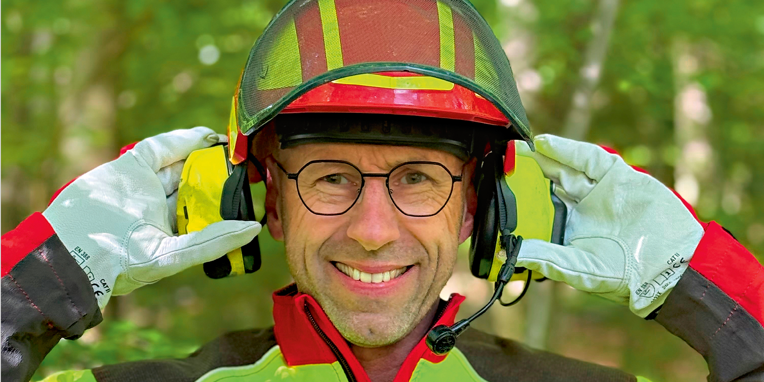 Ein Mann in Persönlicher Schutzausrüstung für Arbeiten im Forst setzt sich lächelnd den Kapsel- Gehörschutz auf. Er trägt einen Helm, Schutzhandschuhe und Jacke. 