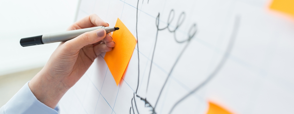 Vergrößerung des Bildes für Flipchart mit der Skizze einer Glühbirne. Darüber klebt ein orangener Klebezettel, der von einer Person beschriftet wird. .