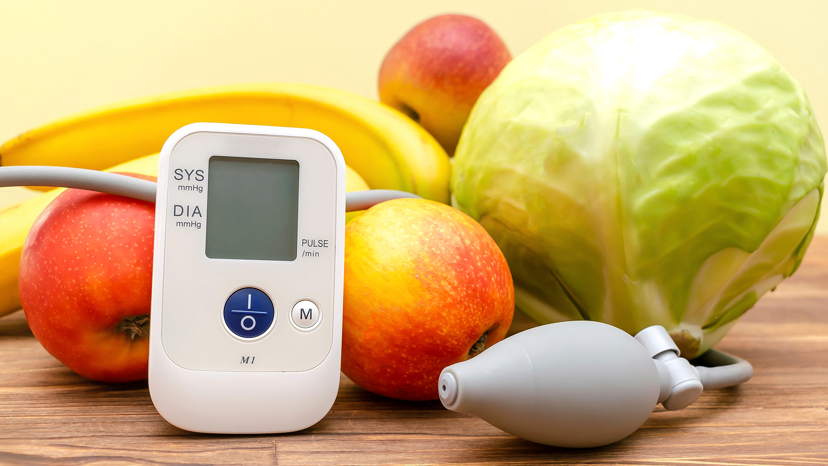 Ein Blutdruckmessgerät liegt auf einem Tisch, umgeben von gesunden Lebensmitteln wie frischem Obst und Gemüse.