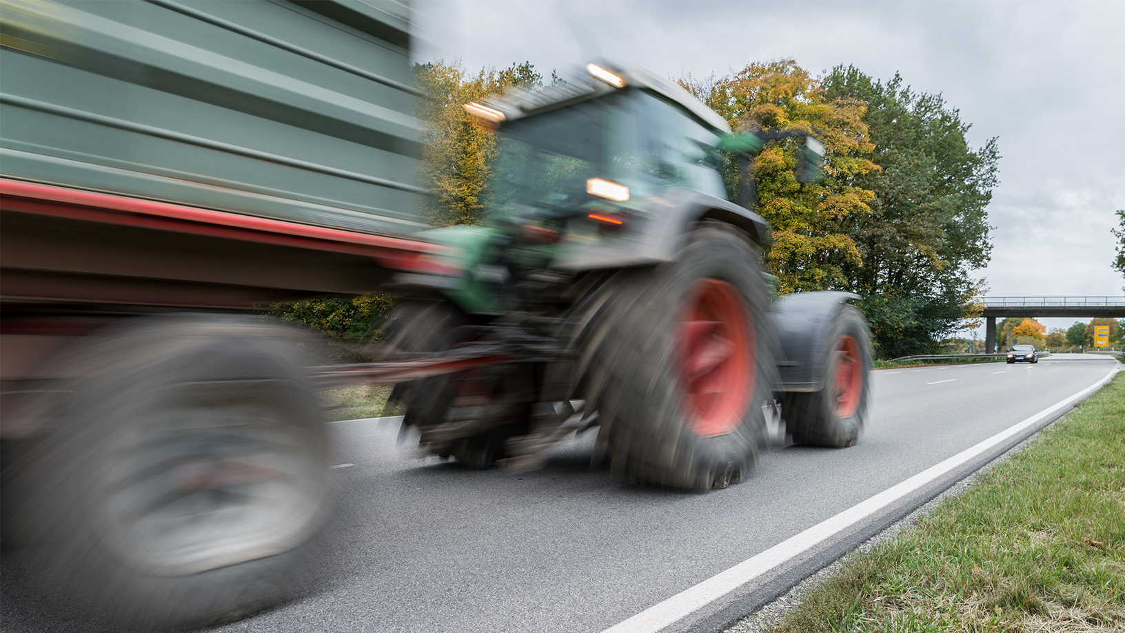 Ein verwaschen wirkender Traktor mit Anhänger fährt auf einer Straße. 
