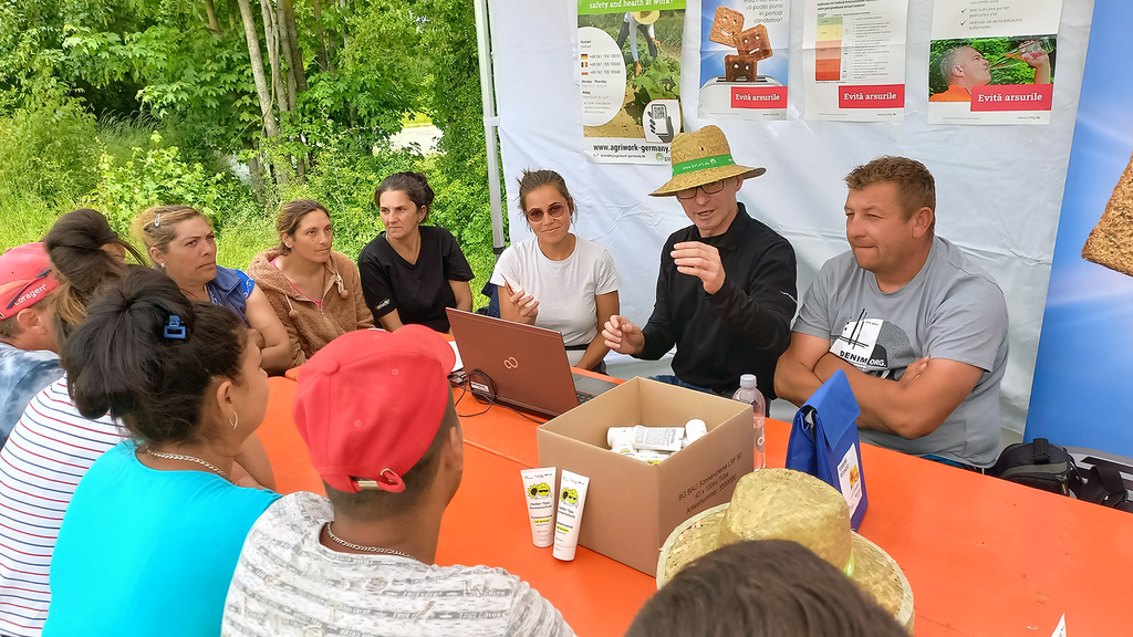 Vergrößerung des Bildes für Eine Gruppe Menschen sitzt in einem Wetterschutzzelt um einen Tisch, auf dem Kisten mit Sonnencreme stehen. An einer Wand des Zeltes hängen Plakate zu Sonnen- und Hitzeschutz. Ein Mann mit Sonnenhut spricht zu den Anwesenden..