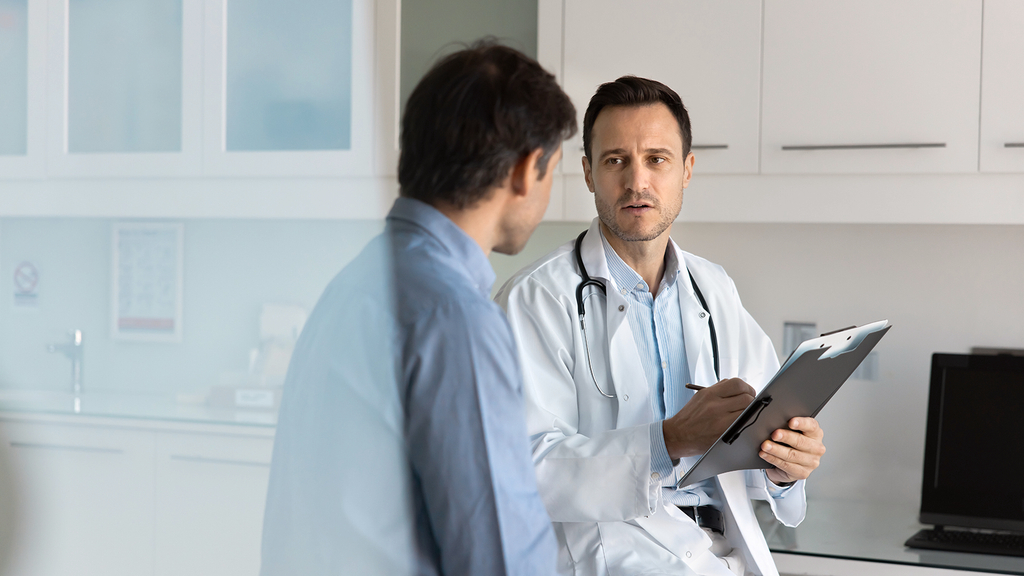 Vergrößerung des Bildes für Ein Arzt, der einen Arztkittel und ein Stetoskoph um den Hals trägt, hält eine Akte in der Hand und spricht dabei mit seinem Patienten. .