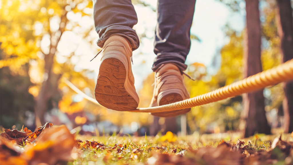 Vergrößerung des Bildes für Das Bild zeigt einen Wald mit Herbstlaub , in dem ein Seil gespannt ist. Auf dem Seil sind zwei Füße in Schuhen zu sehen von jemandem, der darauf balanciert..
