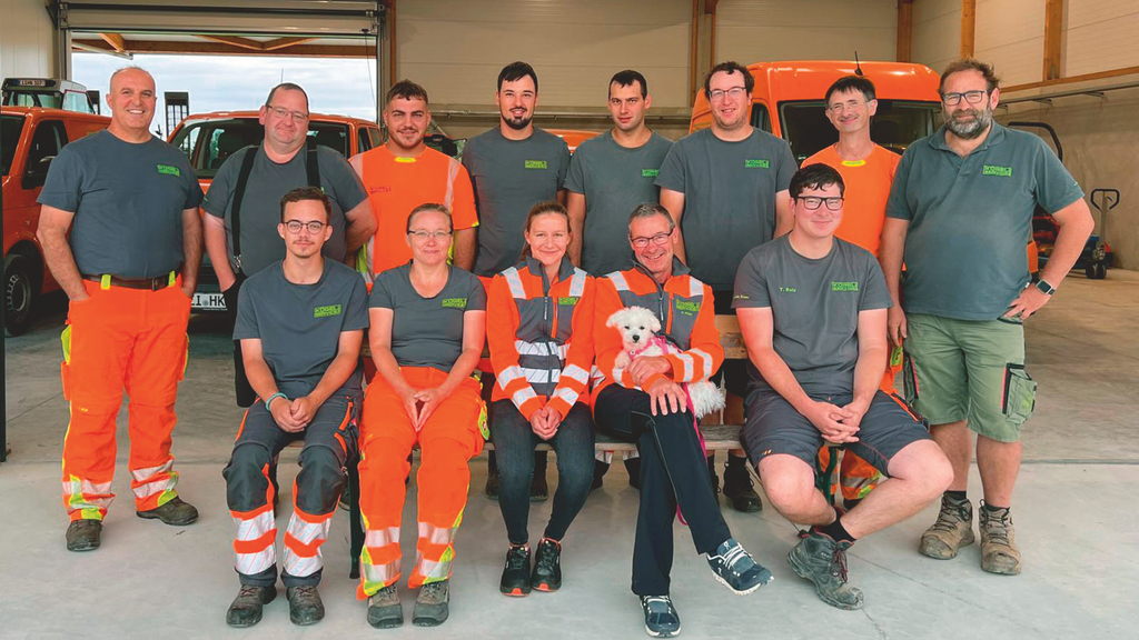 Vergrößerung des Bildes für Gruppenfoto von mehreren Menschen, die in Arbeitskleidung freundlich in die Kamera schauen. Einige haben orange Arbeitshosen mit Reflektoren an. Im Hintergrund sind orange Kleintransporter zu sehen. .