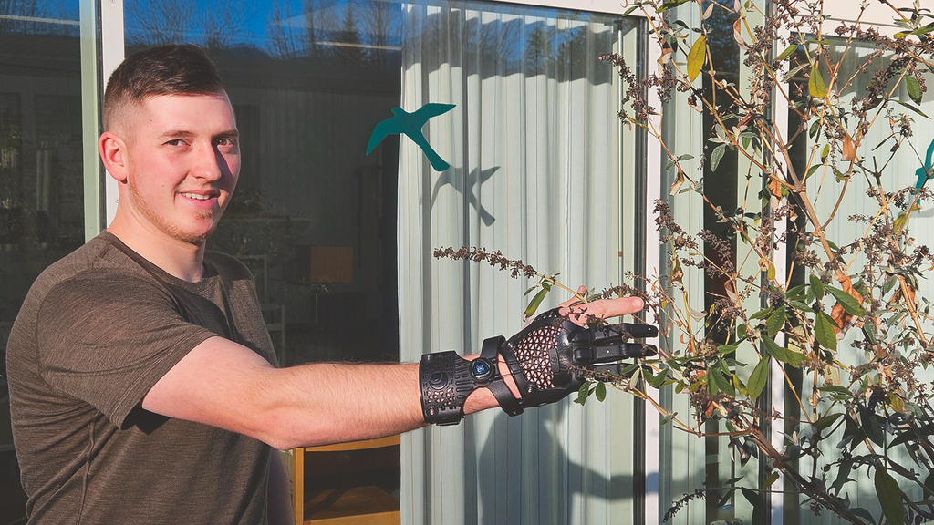 Vergrößerung des Bildes für Ein junger Mann steht im Vorgarten, und fasst mit seiner Teilhandprothese einen Strauch an und schaut dabei in die Kamera..