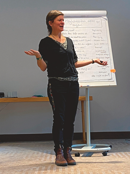 Vergrößerung des Bildes für Eine Frau steht vor einem beschriebenen Flipchart und redet. Dabei hält sie die Unterarme im ungefähren rechten Winkel zum Körper, die Handflächen zeigen nach oben. .