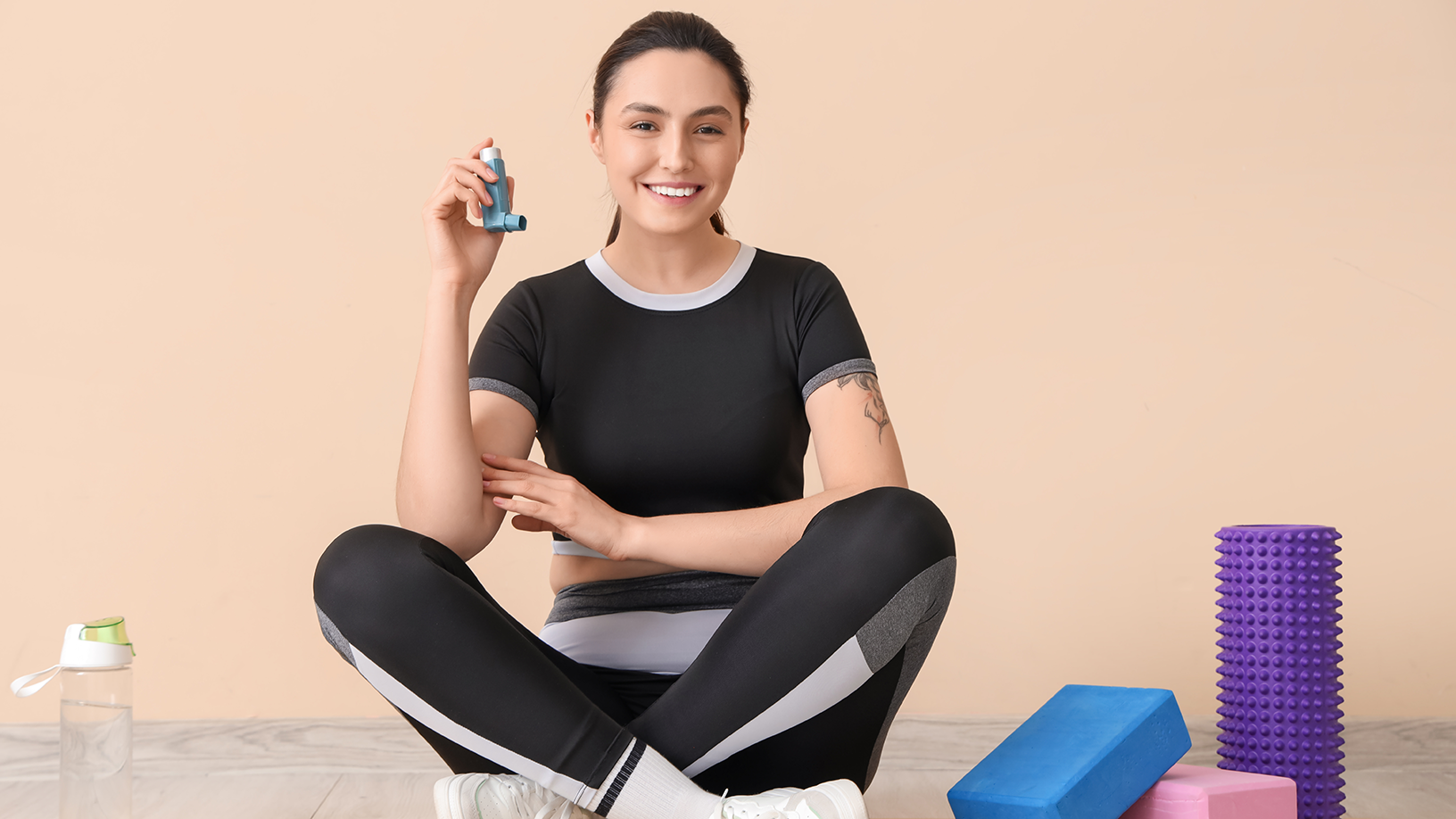 Junge Frau sitzt auf einer Sportmatte, umgeben von kleinen Fitnessgeräten und einer Trinkflasche. Sie hält einen Asthma-Inhalator in der Hand und hält ihn hoch.