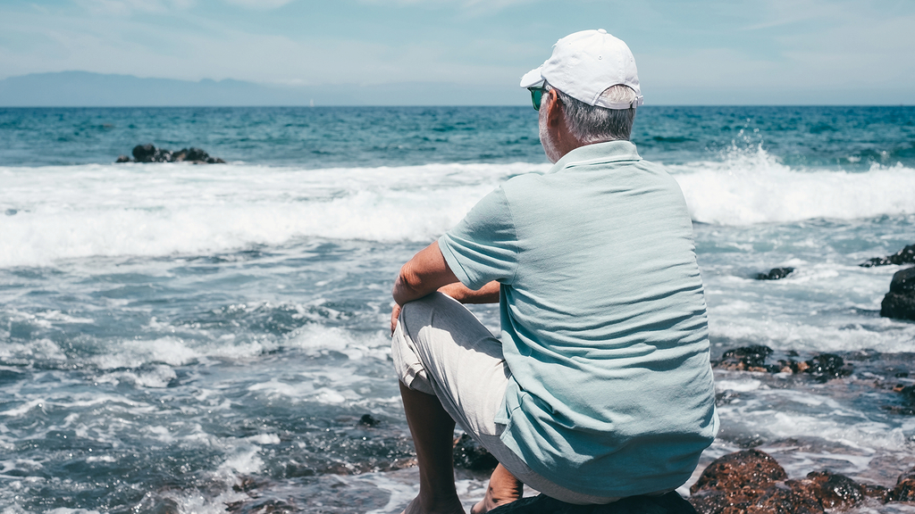 Vergrößerung des Bildes für Ein Mann sitzt auf einem kleinen Fels am Meer und beobachtet die Wellen.