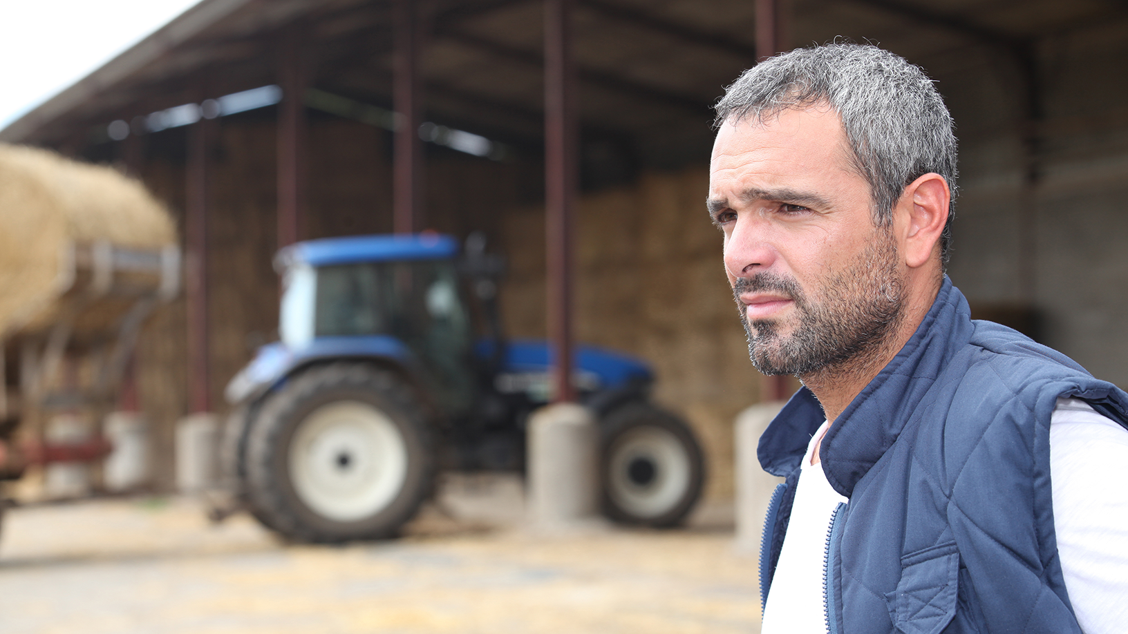 Mann mit einem betrübtem Gesichtsausdruck. Im Hintergrund sieht man ein Hof sowie ein Traktor. 
