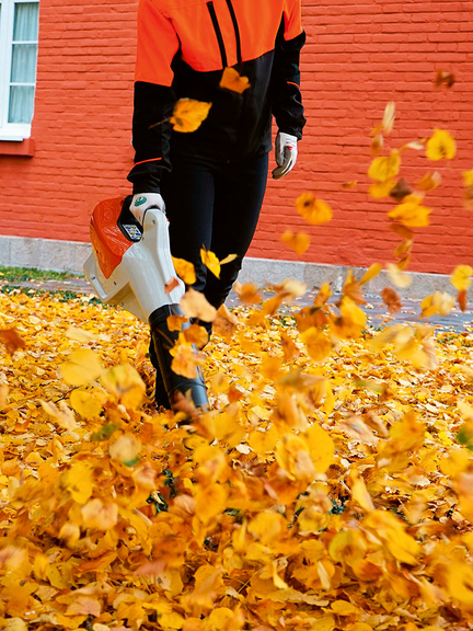Vergrößerung des Bildes für Ein in einer Hand gehaltener Laubbläser pustet buntes Herbstlaut durcheinander.