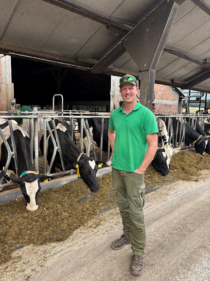 Vergrößerung des Bildes für Ein junger Mann steht lächelnd vor seinem Rinderstall. Die Rinder sind im Fangfressgitter und haben Futter bekommen.